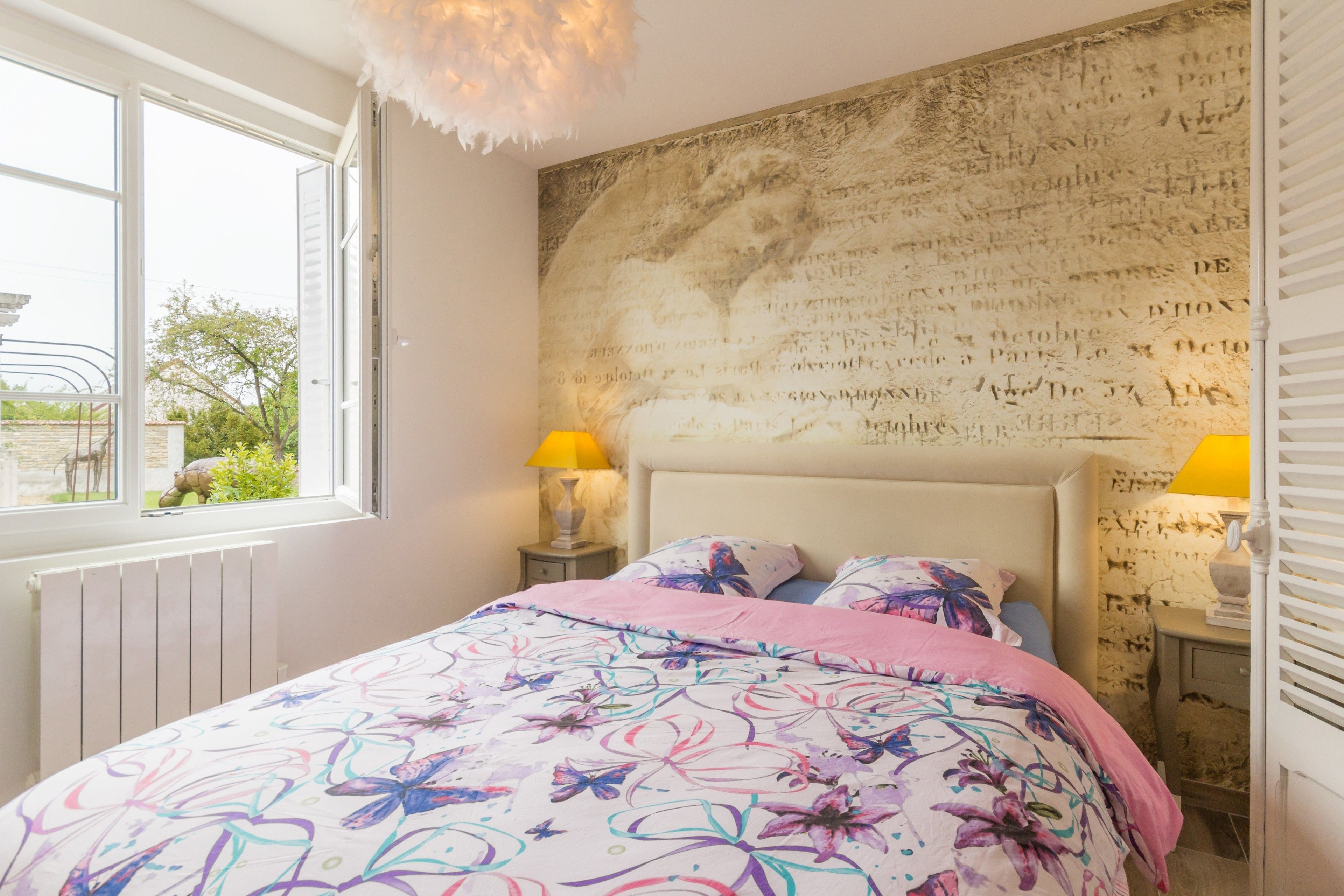 Un dormitorio con un ventanal grande, una cama con cubrecama floral, una pared con letras decorativas y dos lámparas de noche.