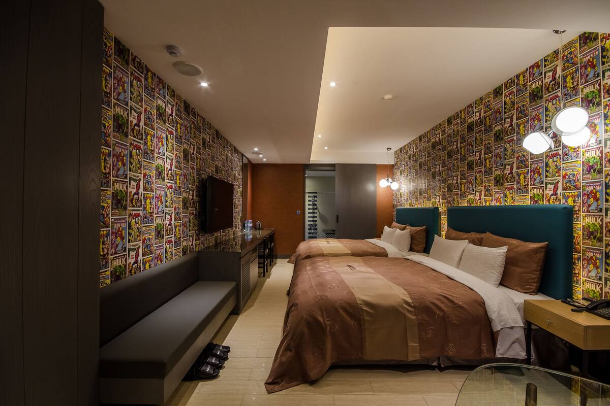 Una habitación de hotel moderna con una cama grande, un banco y un escritorio. Las paredes están decoradas con papel tapiz estilo cómic.