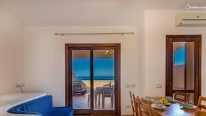 A living room with a sofa, a dining table with chairs, and a view of the sea through a glass door. - Casa Rosa Quattro (Trinità d'Agultu e Vignola)