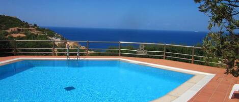 Una piscina rettangolare con ringhiera metallica, circondata da piastrelle in terracotta, con vista sul paesaggio costiero.