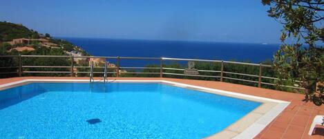 A rectangular swimming pool with a metal railing, surrounded by terracotta tiles, and overlooking a coastal view.
