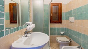 A bathroom with a sink, toilet, and bidet, tiled in green and beige, with a mirror and a wooden cabinet above the toilet. - Casa Rosa Due (Trinità d'Agultu e Vignola)