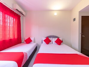 A hotel room with two beds, each with red pillows and white bedding. There is an air conditioning unit on the wall, a door on the right, and a small window with a view of the outside.