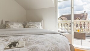 Une chambre avec un grand lit, une fenêtre donnant sur des immeubles et un balcon avec une balustrade.
