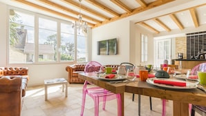 Un vaste salon doté d’un plafond en bois, de grandes fenêtres, d’un canapé et d’une table à manger agrémentée de chaises roses.