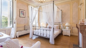 Un lit à baldaquin à quatre poteaux, avec une table de chevet et une lampe, un banc avec un coussin, et une vue sur la verdure à l ’extérieur.