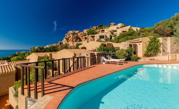 A swimming pool with a curved edge, a wooden deck, and a view of a hillside with buildings. - Villa Violetta 1 Bianca (Trinità d'Agultu e Vignola)