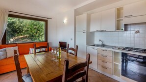 A modern kitchen with white cabinets, a gas stove, and a sink. There is a dining area with a wooden table and chairs, and a sofa with orange cushions. - Villa Violetta 1 Bianca (Trinità d'Agultu e Vignola)