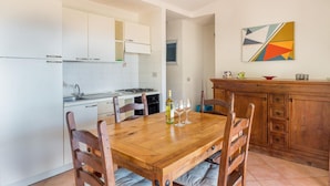 A compact kitchen with white cabinets, a wooden dining table, and a wooden cabinet with drawers. - Villa Violetta 1 Bianca (Trinità d'Agultu e Vignola)
