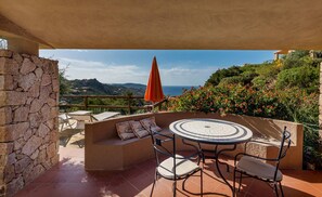 Une terrasse extérieure avec un mur en pierre, une table ronde entourée de chaises, un parasol rouge et une vue sur la mer et les collines.