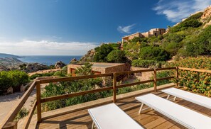 A wooden deck with white lounge chairs overlooking a scenic coastal landscape with buildings on a hillside. - Villa Violetta 2 Celeste (Trinità d'Agultu e Vignola)