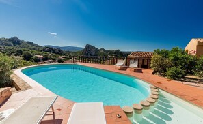 A swimming pool with a curved edge, surrounded by a tiled deck and a stone wall. There are two lounge chairs and a small structure with a tiled roof. - Villa Violetta 2 Celeste (Trinità d'Agultu e Vignola)