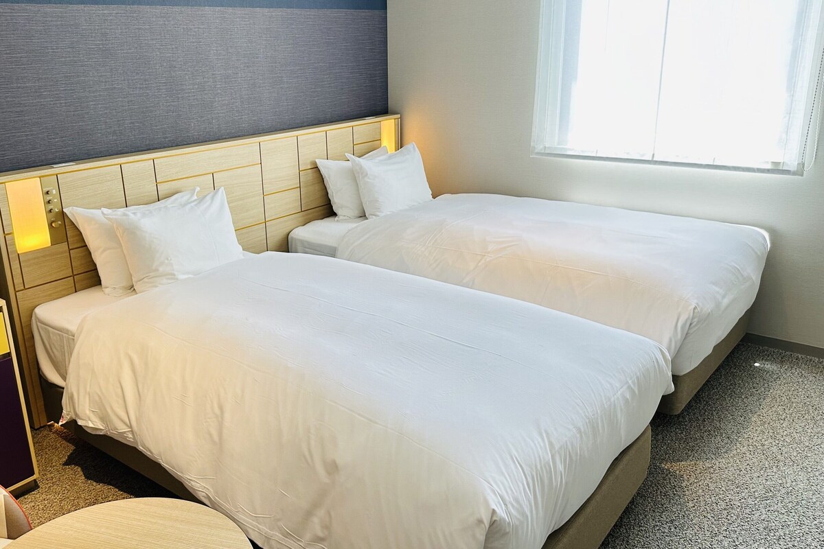 A hotel room with two beds, a wooden headboard, a small desk, and a window with curtains.