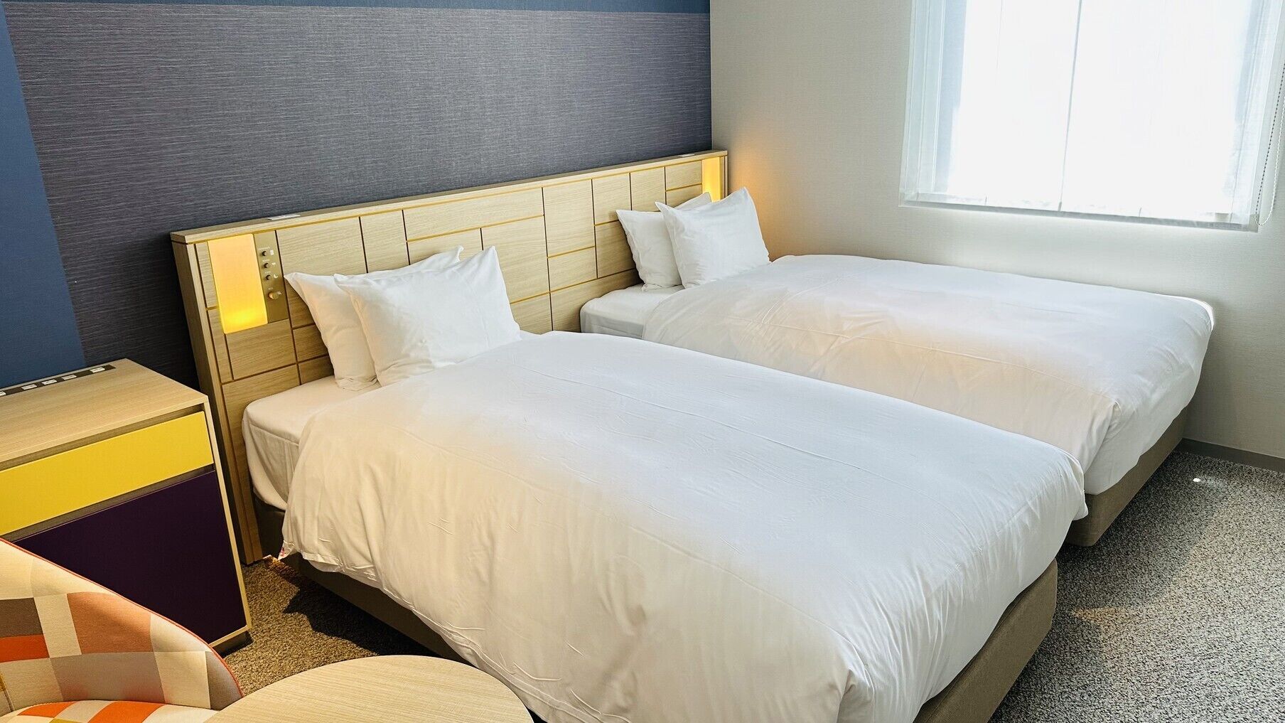 A hotel room with two beds, a wooden headboard, a small desk, and a window with daylight.