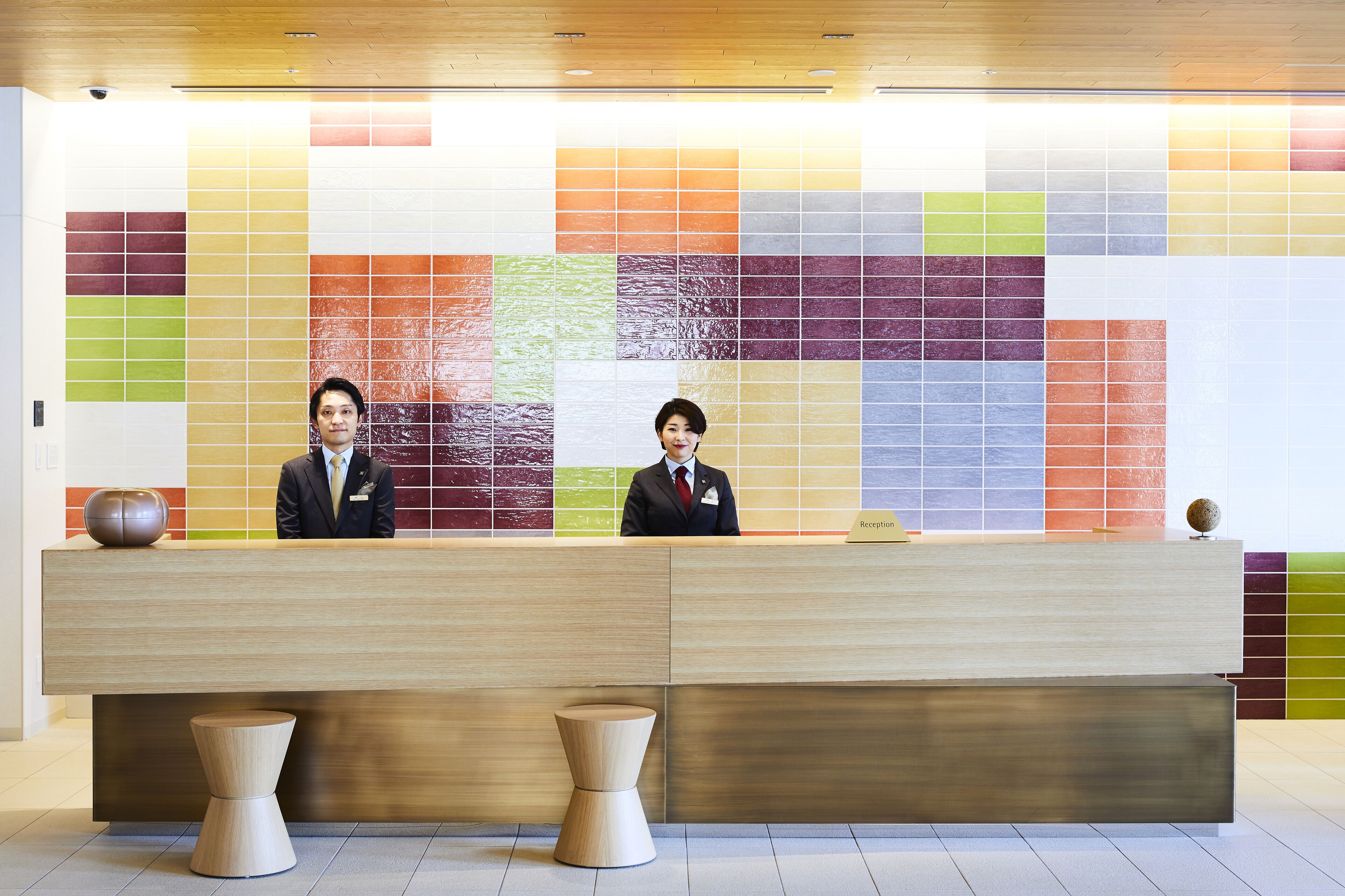A reception desk with two staff members, a tiled wall with a multicolored pattern, and two wooden stools.