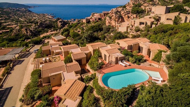 A resort with a swimming pool surrounded by terracotta-roofed buildings, set against a backdrop of a coastal landscape.