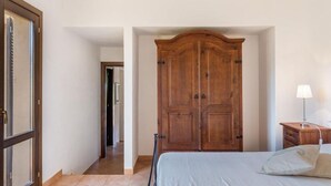 A bedroom with a bed, a wooden wardrobe, a nightstand with a lamp, and a door leading to another room.