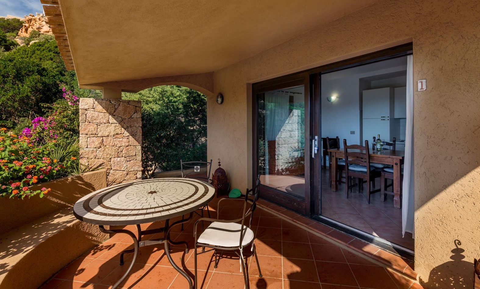 A patio with a round table and chairs, a stone wall, and a view of the interior through a glass door.