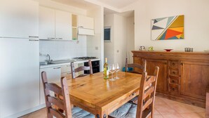 A kitchen with white cabinets, a wooden dining table with chairs, and a painting on the wall. - Villa Violetta 3 Verde (Trinità d'Agultu e Vignola)