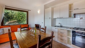 A modern kitchen with white cabinets and a tiled backsplash, a dining area with a wooden table and chairs, and a large window with a view of greenery. - Villa Violetta 3 Verde (Trinità d'Agultu e Vignola)