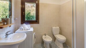 A bathroom with a sink, bidet, toilet, and a window with a view of greenery. - Villa Violetta 5 Rossa (Trinità d'Agultu e Vignola)