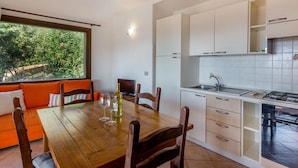 A modern kitchen with white cabinets, a wooden dining table, and a view of greenery outside. - Villa Violetta 5 Rossa (Trinità d'Agultu e Vignola)