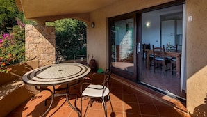An outdoor patio with a round table and chairs, a stone wall, and a view into a dining area through a glass door. - Villa Violetta 5 Rossa (Trinità d'Agultu e Vignola)