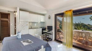 A modern kitchen with white cabinets, a dining table with a blue tablecloth, and a balcony with a yellow curtain. - Villetta Ginepro Rosso (Trinità d'Agultu e Vignola)