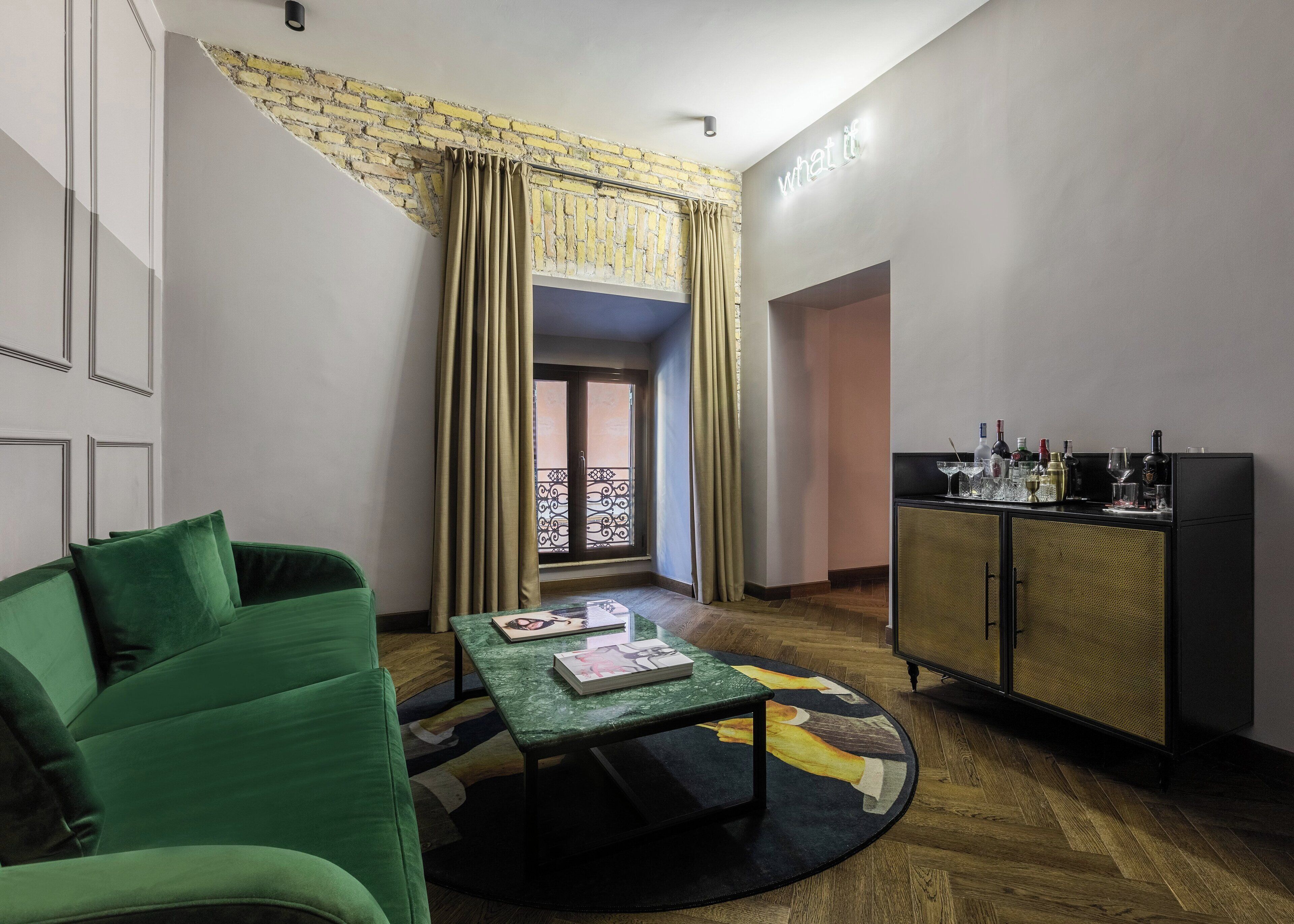 A room with a green sofa, a small table with a book, a cabinet with bottles, and a window with curtains.