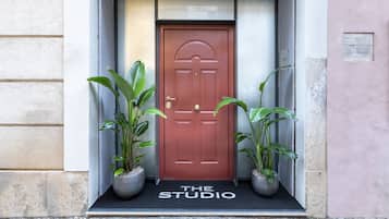A red door with a doormat reading "THE STUDIO" and two potted plants on either side.