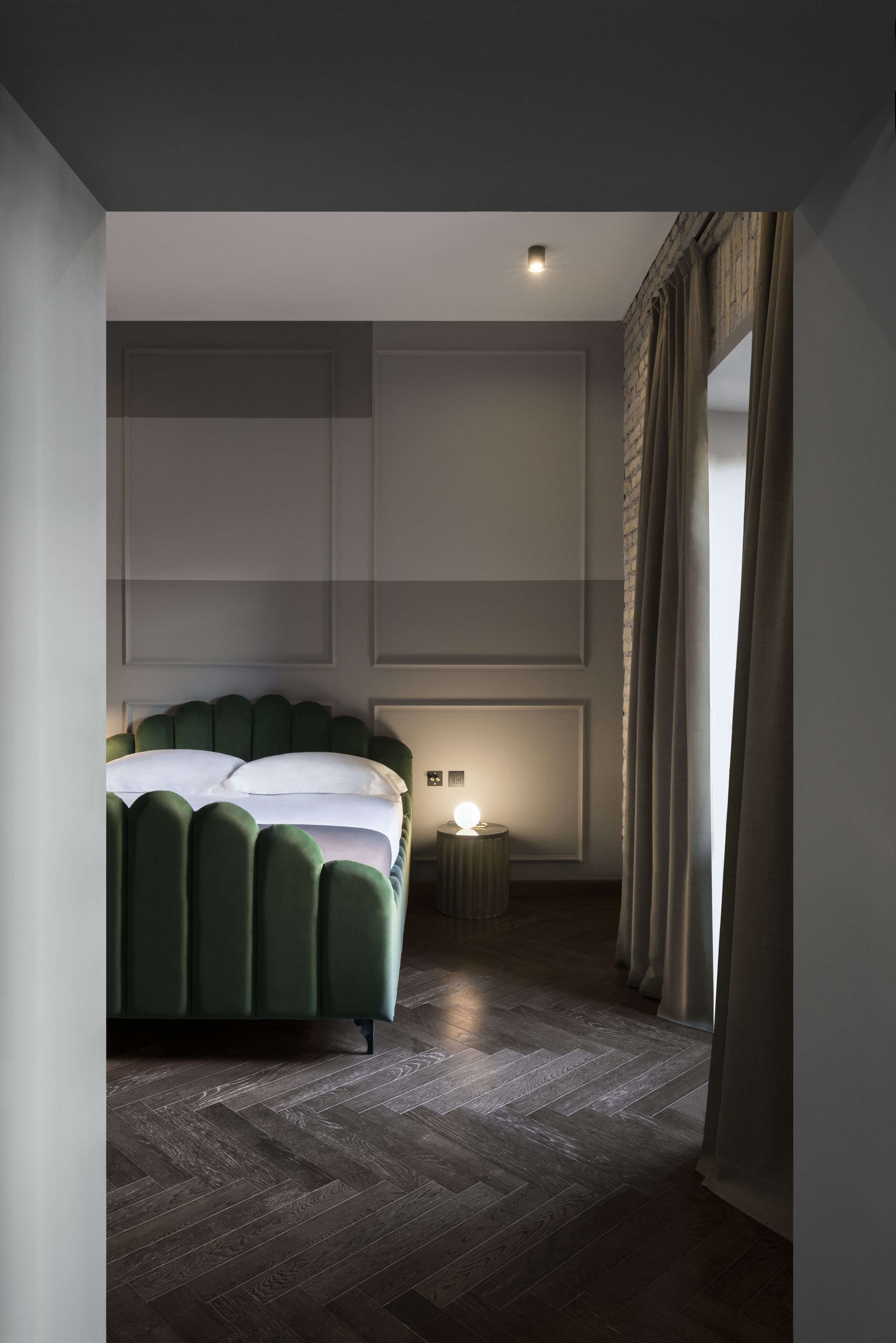 Habitación de hotel moderna con una cama tapizada de verde, una mesita de noche con lámpara y un ventanal con cortinas.