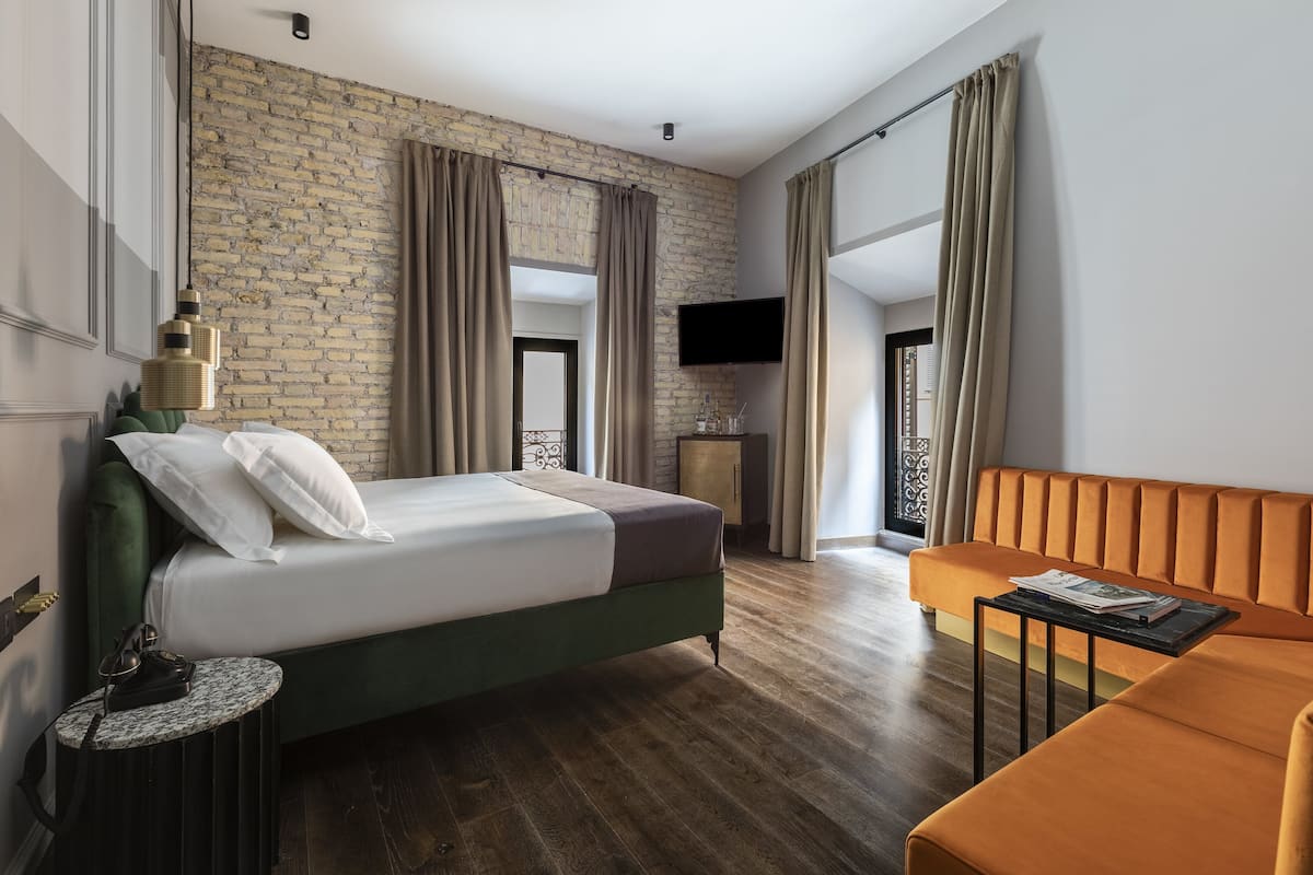 A modern hotel room with a brick wall, a bed with white and gray bedding, a green upholstered headboard, a small round table with a lamp, a sofa, and a flat-screen TV mounted on the wall.