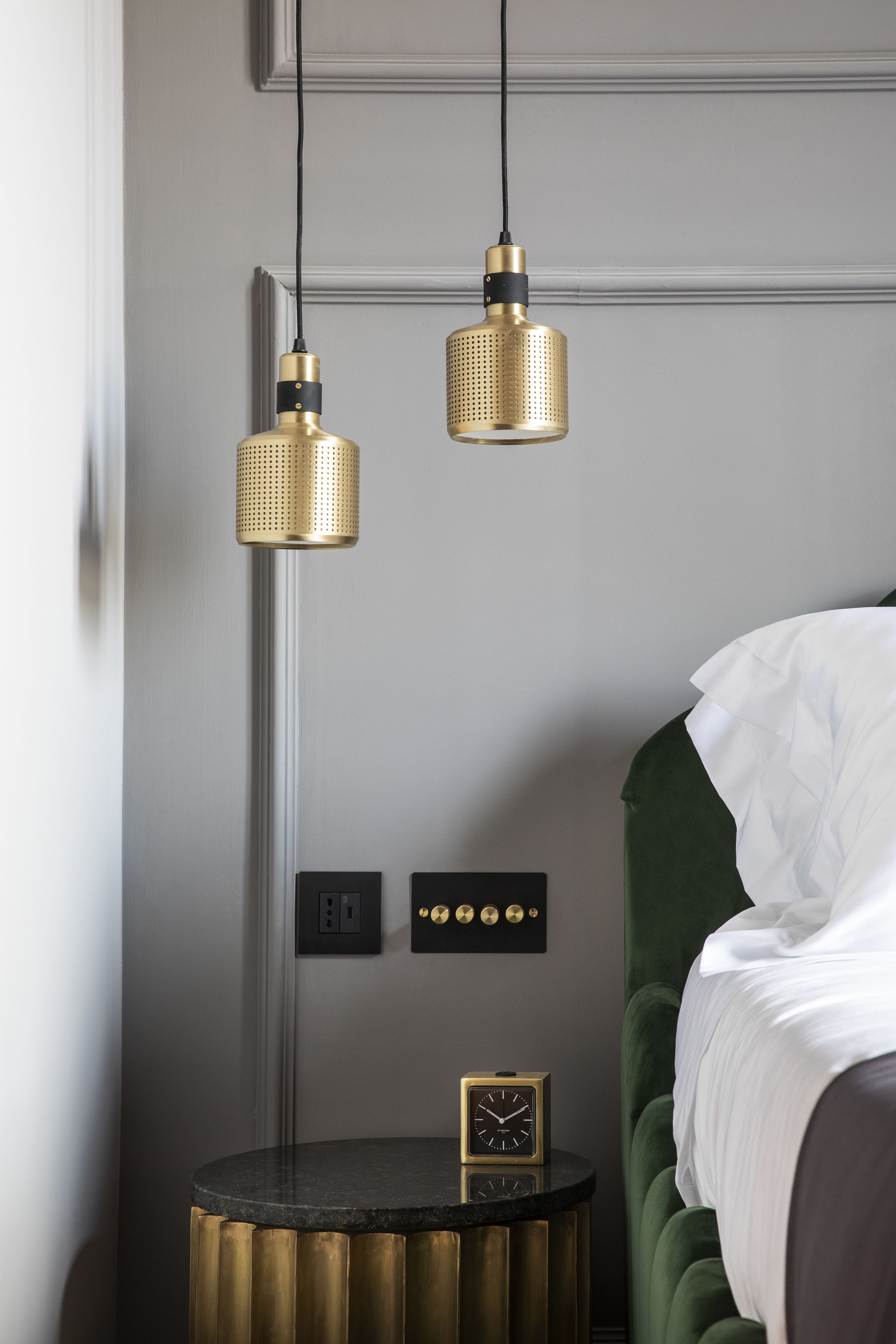 A bedroom with a green upholstered bed, a round black side table with a gold base, and two hanging gold pendant lights.