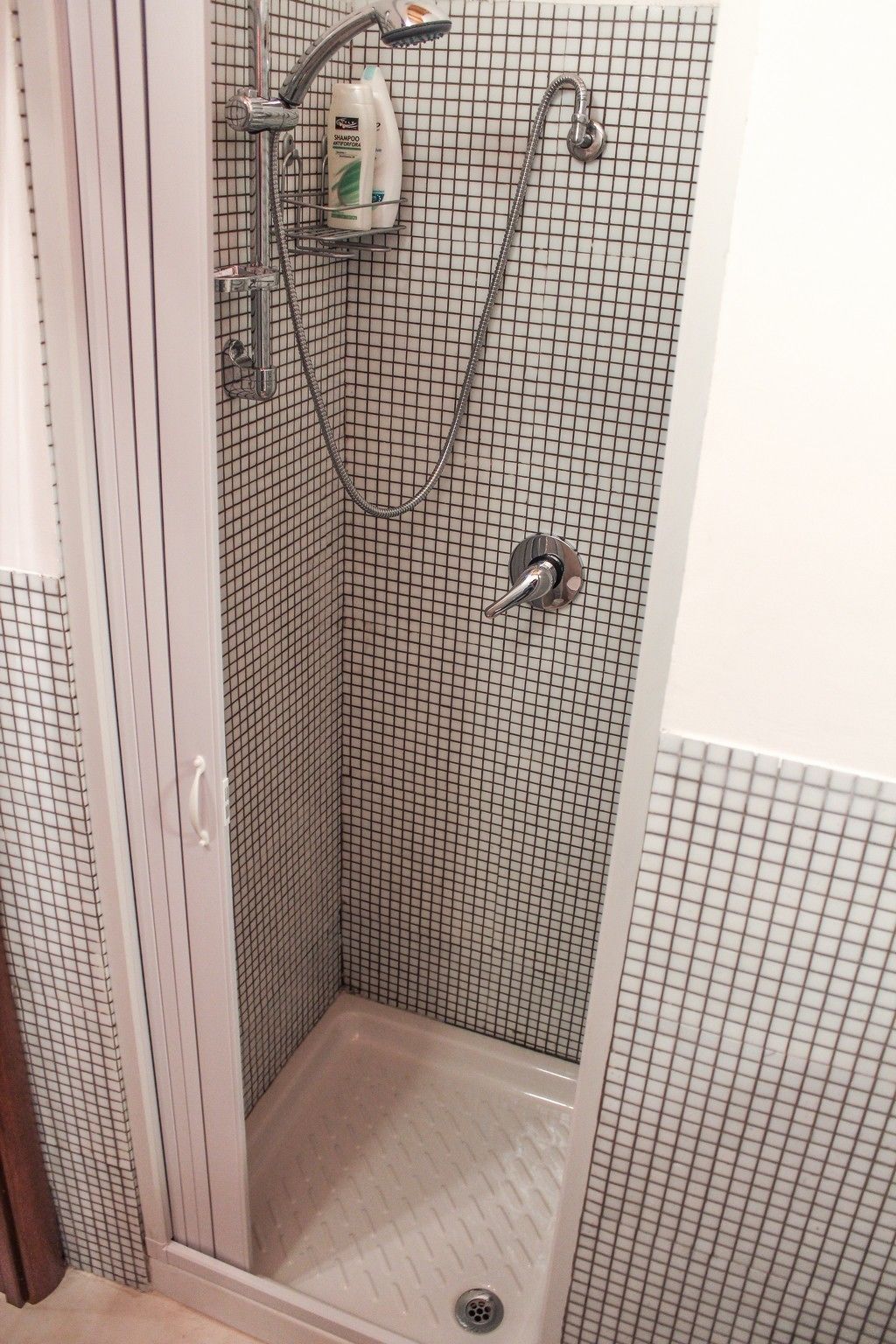 Un espacio de ducha pequeño y cerrado con un revestimiento de pared de azulejos en patrón de cuadrícula, un cabezal de ducha manual y un accesorio de ducha manual.