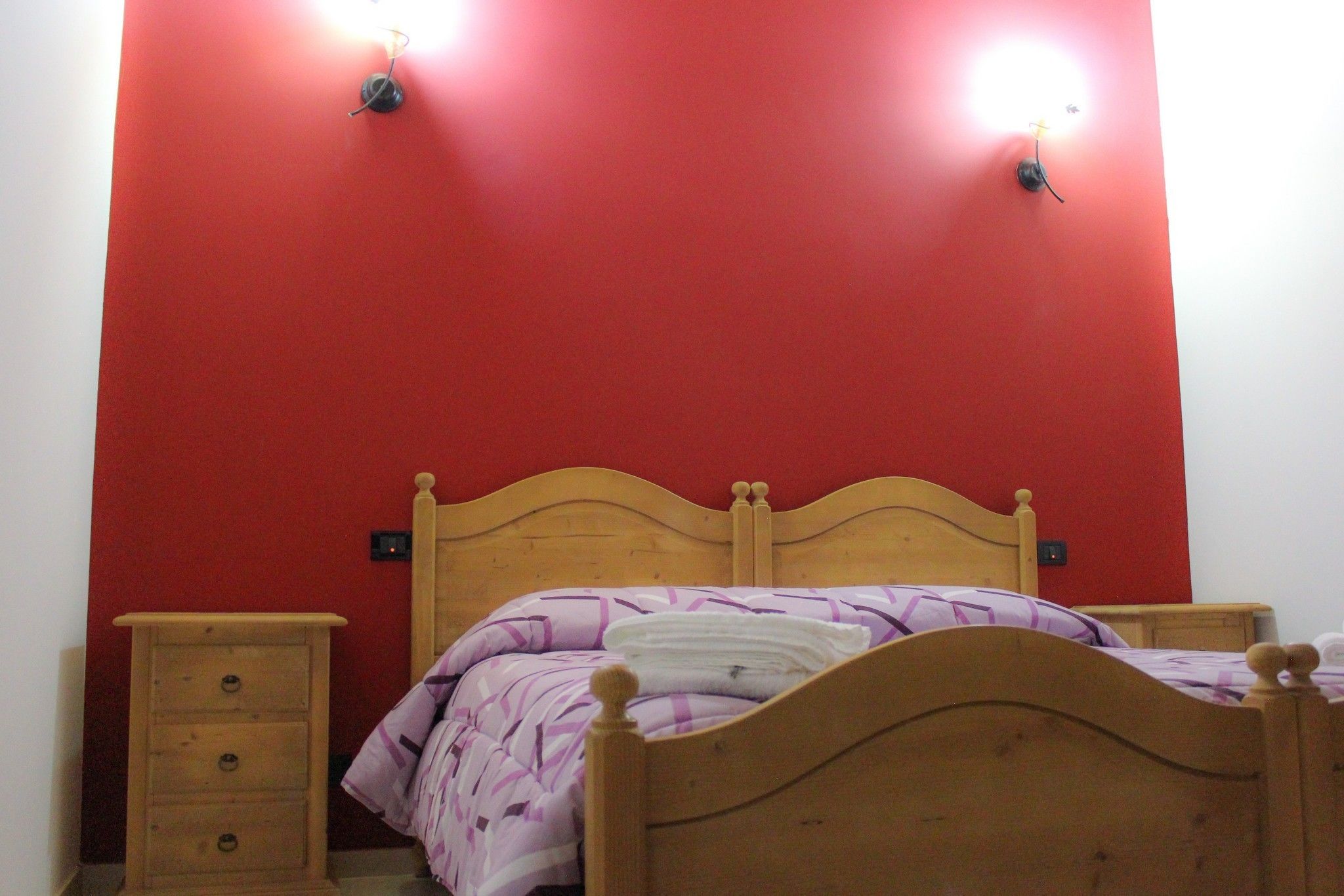 Una cama individual con cabecera de madera en habitación con paredes rojas y dos lámparas fijadas a la pared.