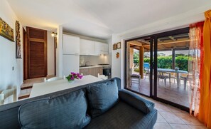 A modern kitchen with white cabinets, a dining table with a vase of flowers, and a sliding glass door leading to an outdoor patio area. - Villetta Ginepro Verde (Trinità d'Agultu e Vignola)