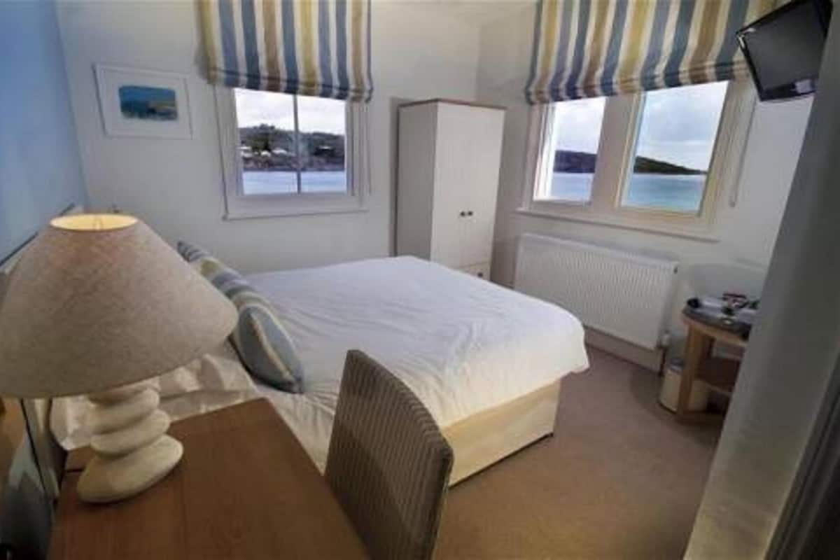 A bedroom with a bed, a desk with a lamp, a chair, a wardrobe, and a view of the sea through windows.