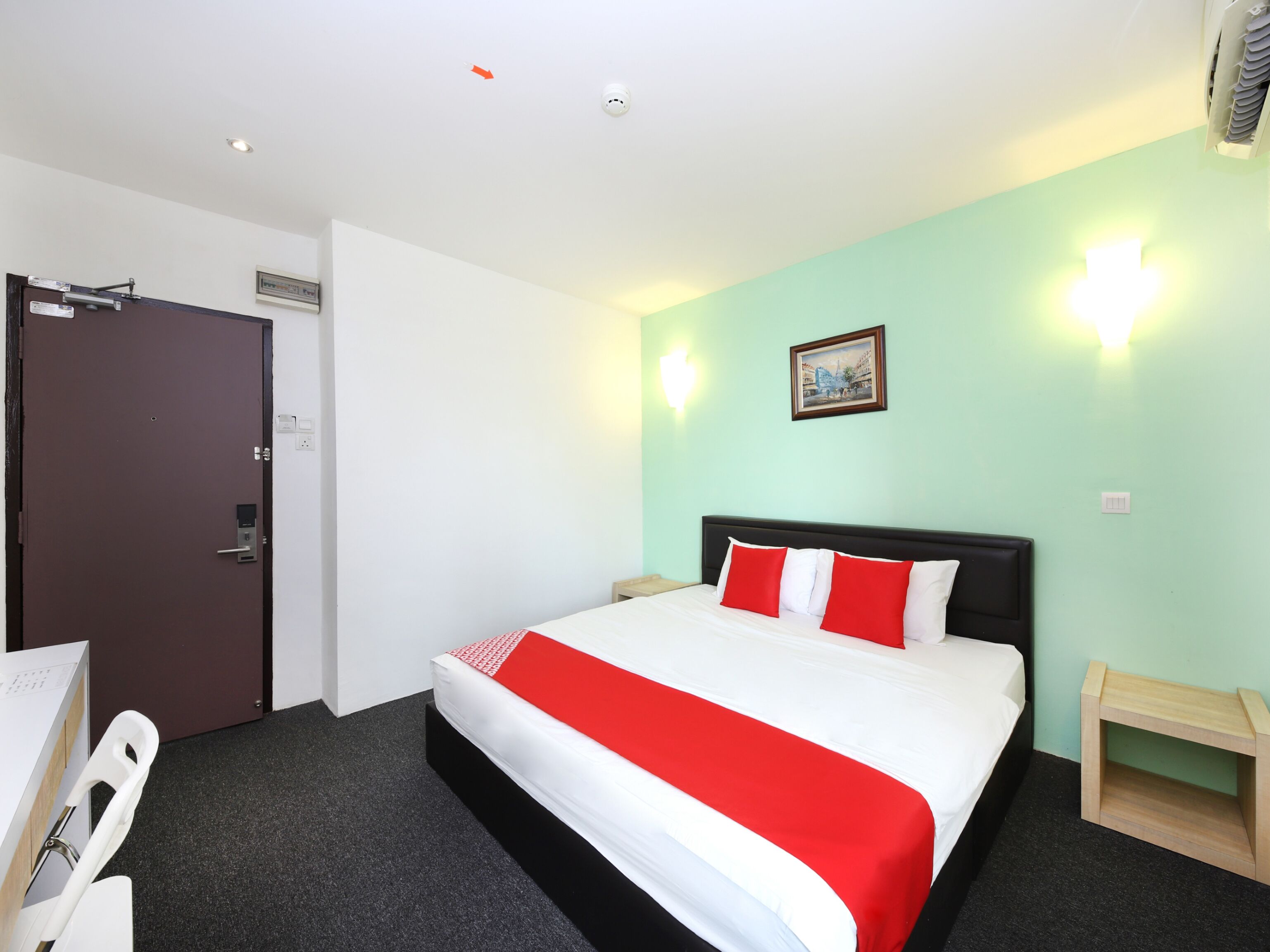 Una habitación de hotel con cama, escritorio, silla, puerta y lámpara fijada a la pared.