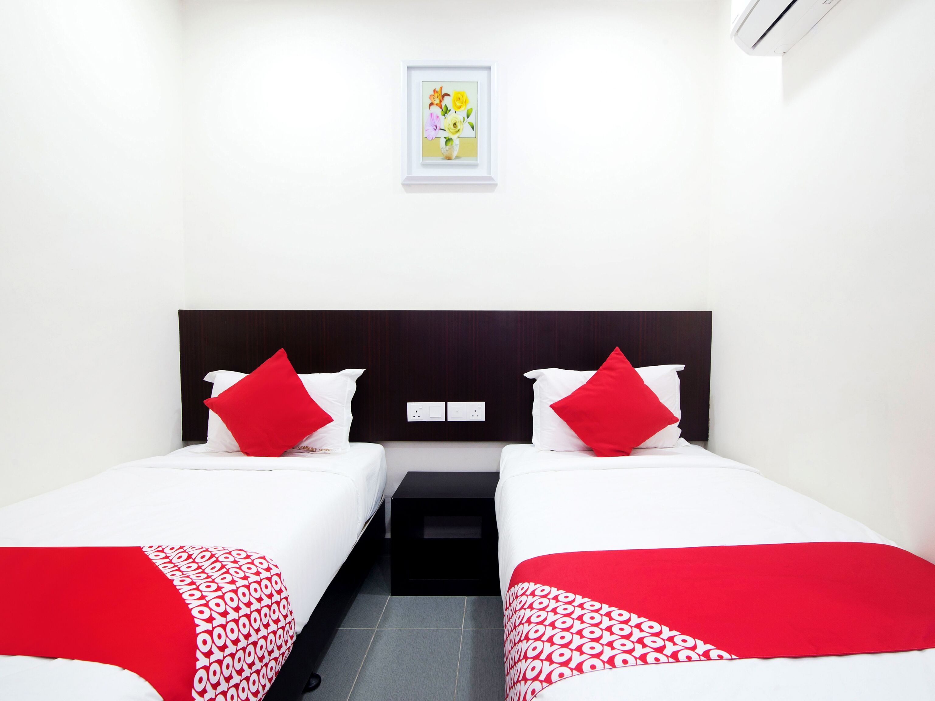 A hotel room with two beds, each with white bedding and red accents, a small bedside table, a framed picture on the wall, and a white air conditioning unit.