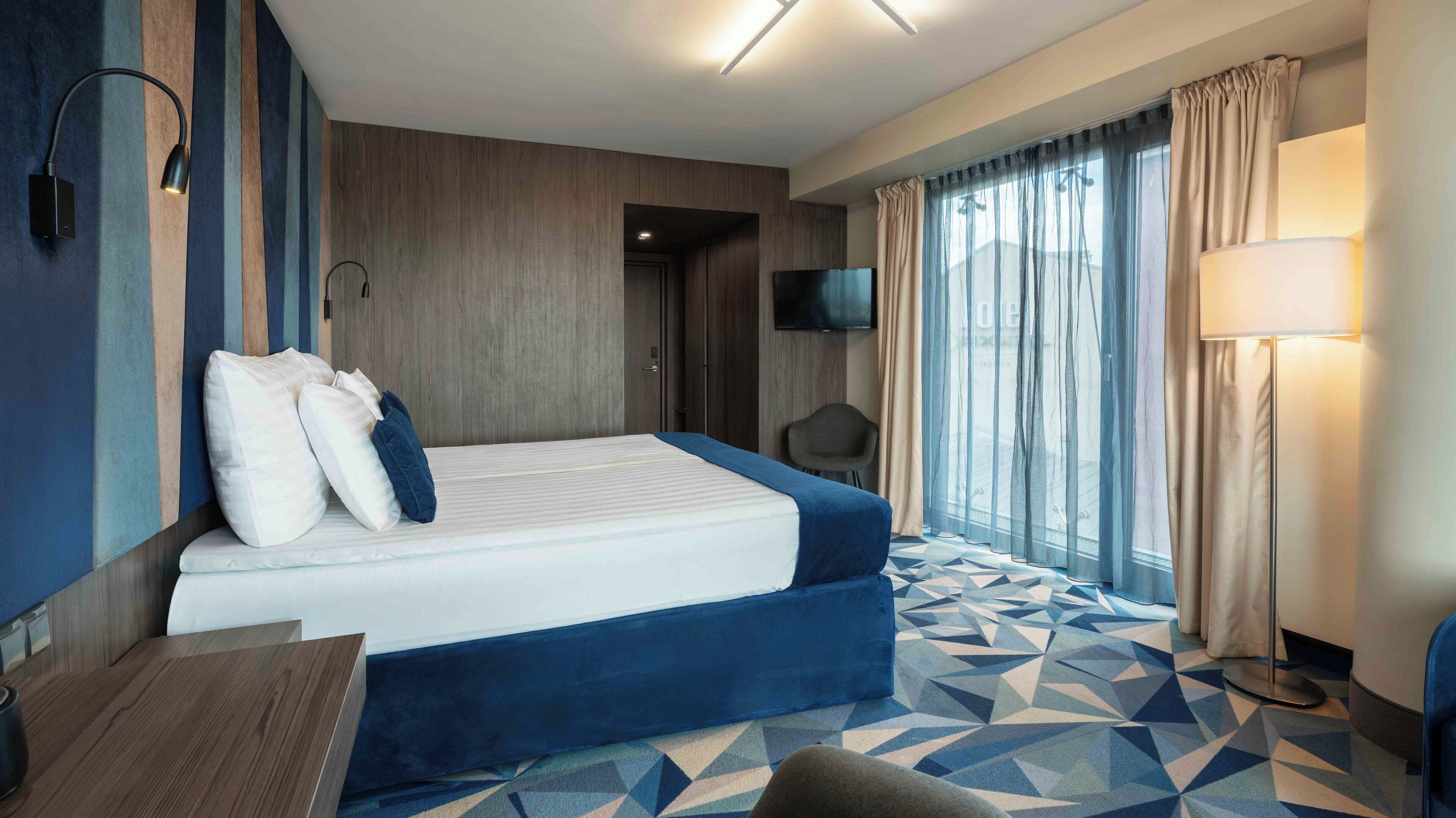 Habitación de hotel con cama, escritorio, silla, televisión y ventana con cortinas.