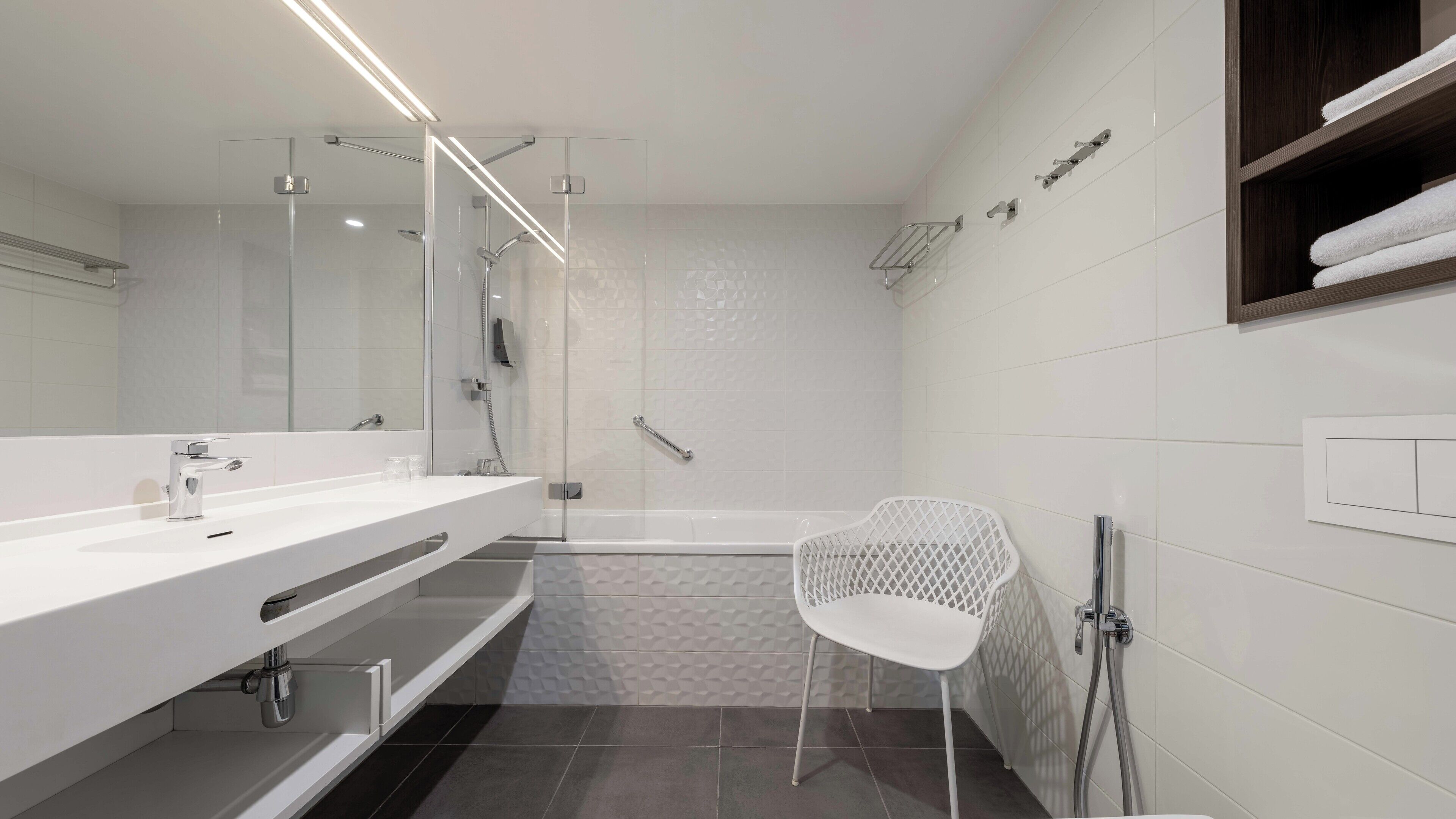 A modern bathroom with a large mirror, a white vanity, a shower with a glass door, and a white chair.