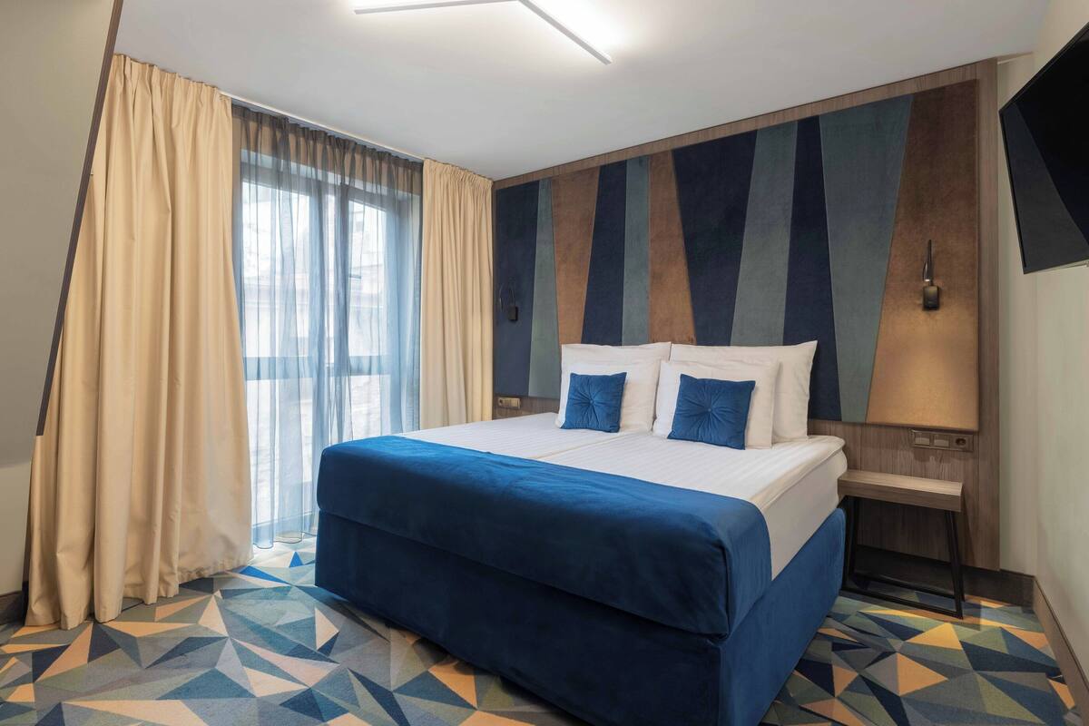 A hotel room with a bed, a TV, a desk, and a window with curtains.