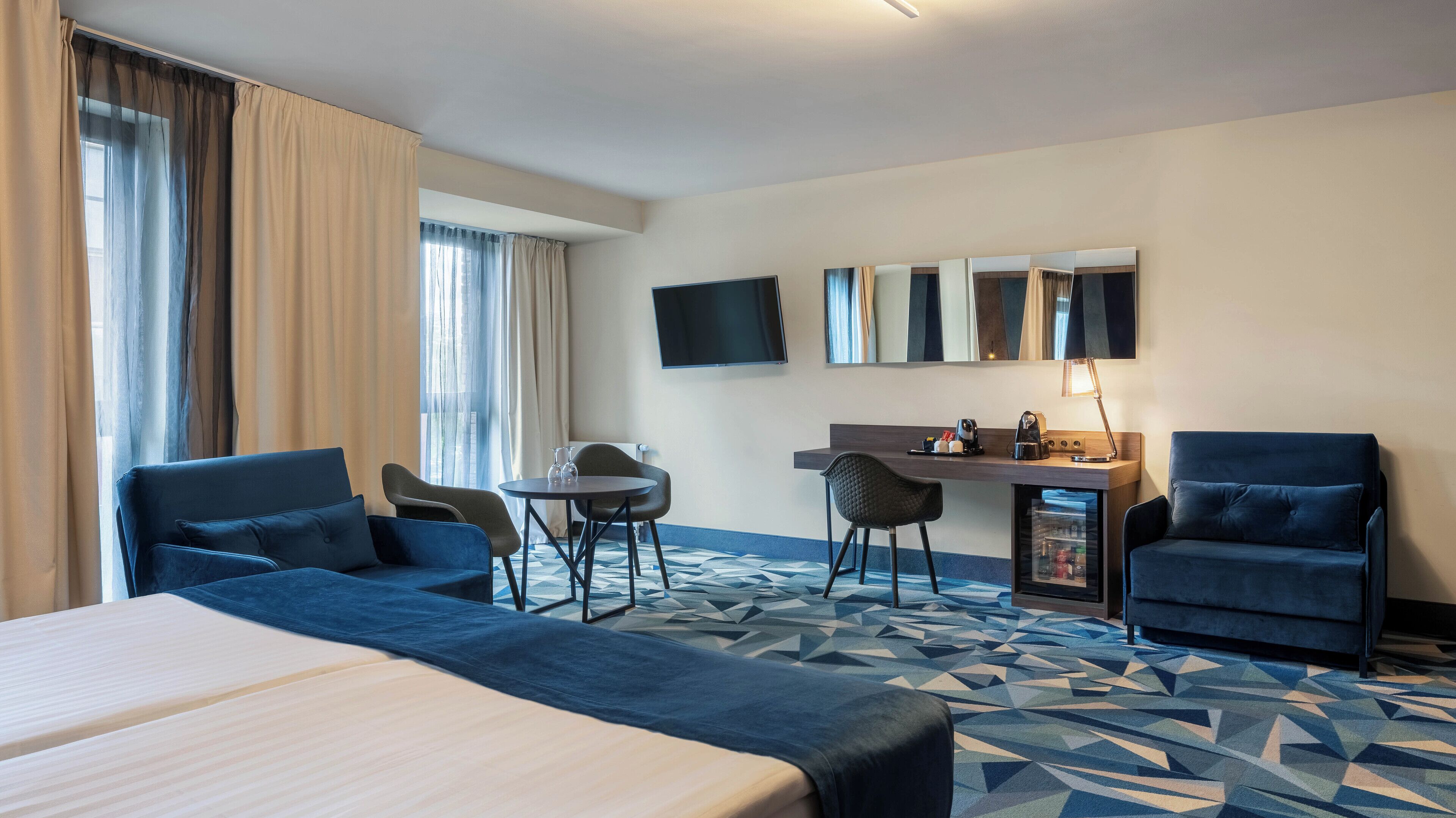 A hotel room with a large bed, two armchairs, a desk with a computer, a flat-screen TV, and a minibar.
