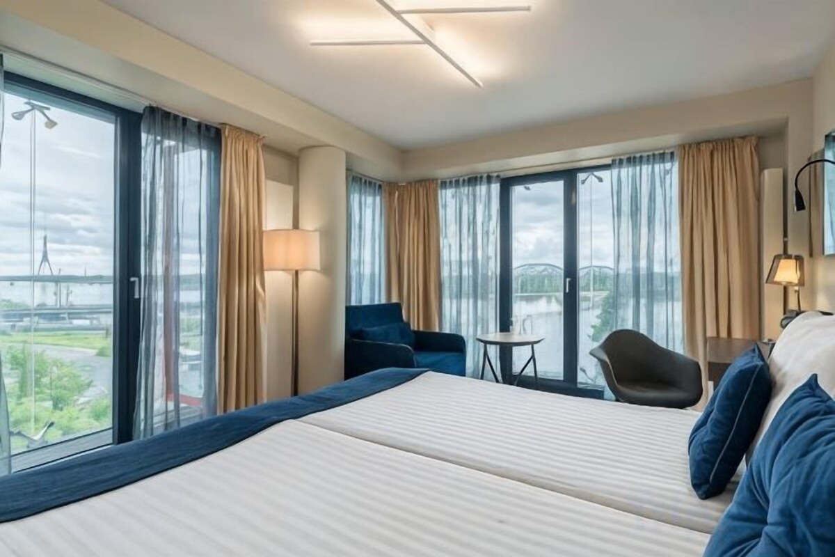 A hotel room with a large bed, a chair, a small table, and a view of a bridge.