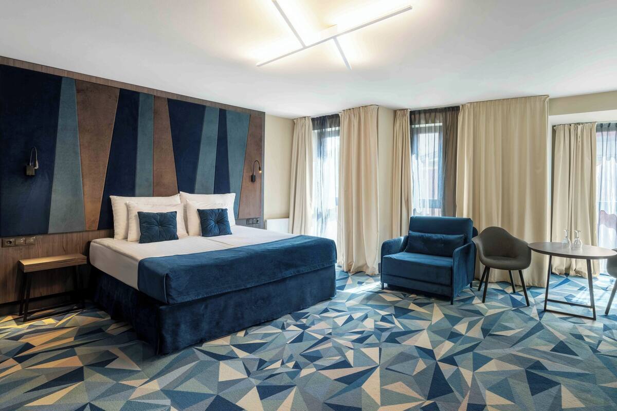 A hotel room with a large bed, two armchairs, a small table, and a geometric patterned carpet.