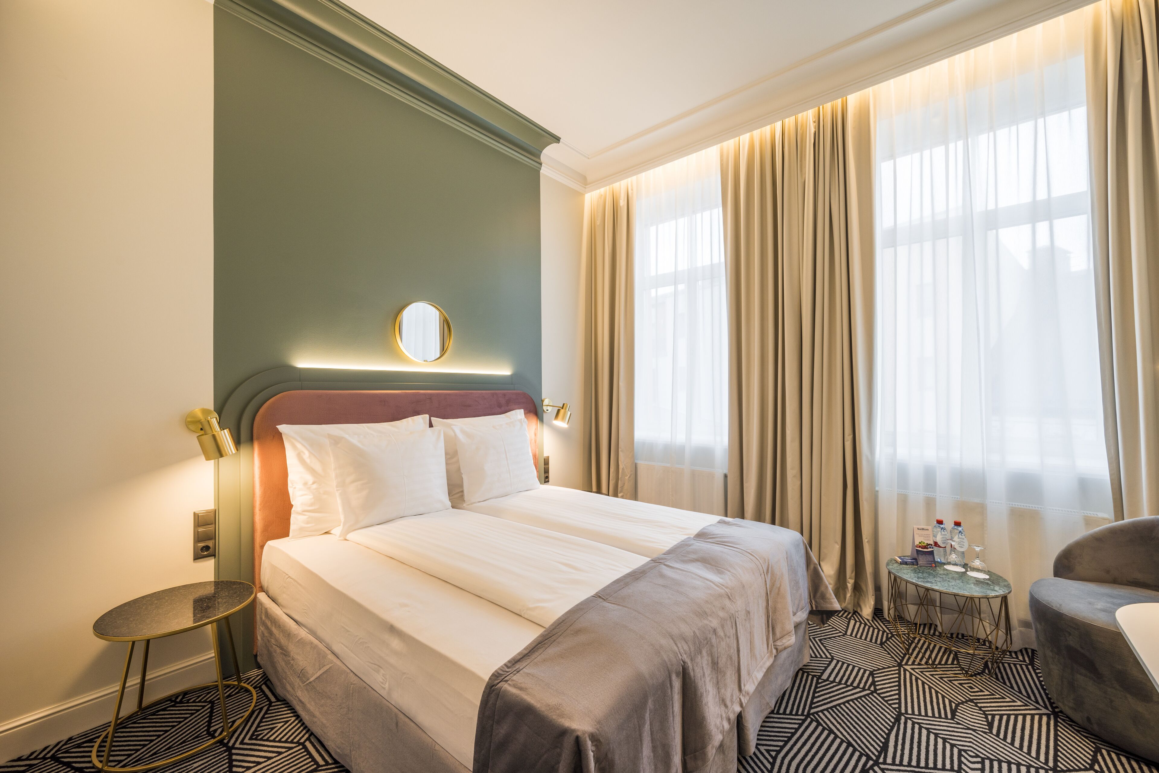 A hotel room with a large bed, a round mirror above the headboard, a small round table, and a chair.