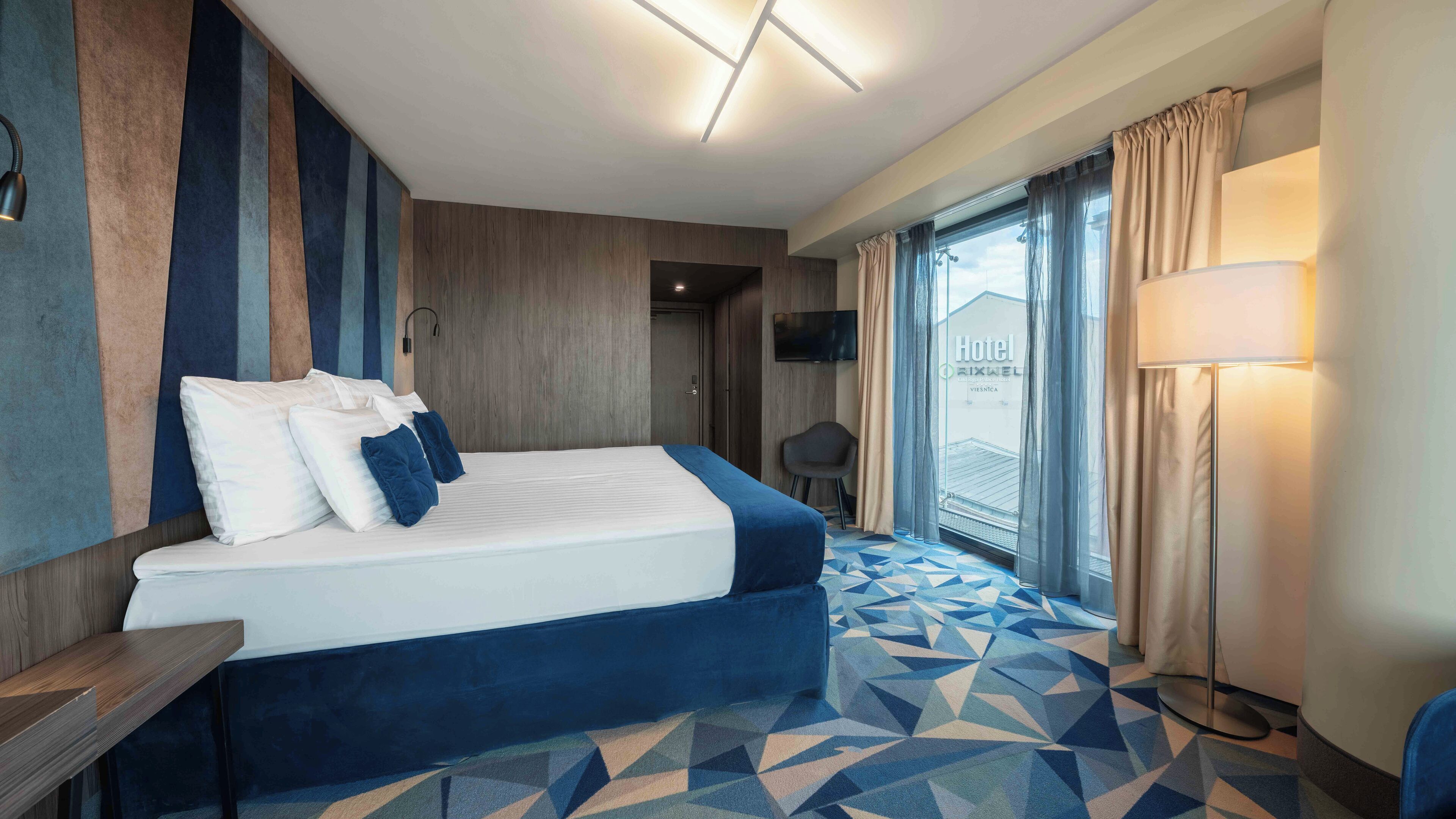 Une chambre d’hôtel avec un lit, un bureau, une chaise, une télévision et une fenêtre avec des rideaux.
