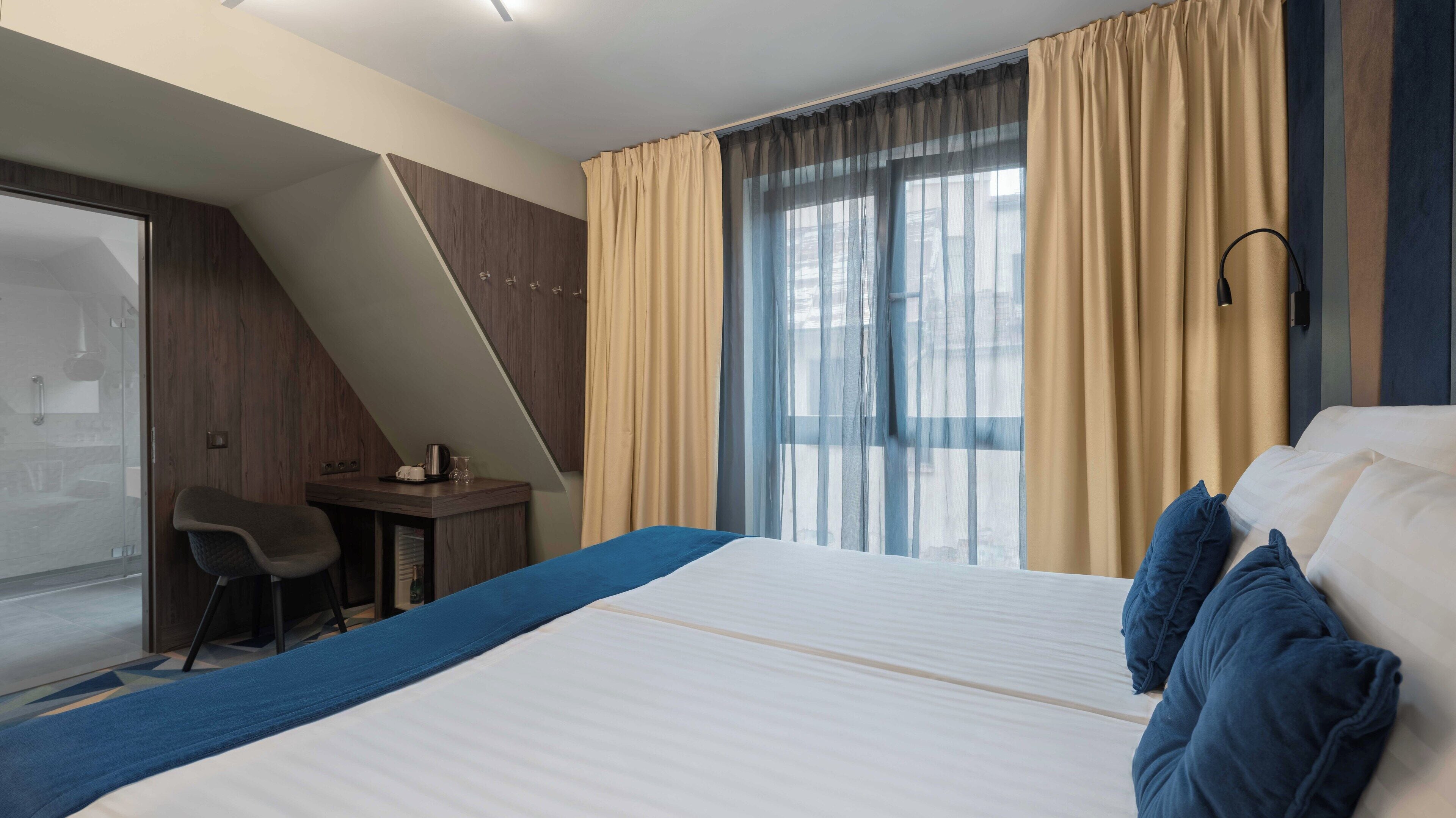 A hotel room with a bed, a desk, and a chair. There is a window with curtains and a view of the outside.