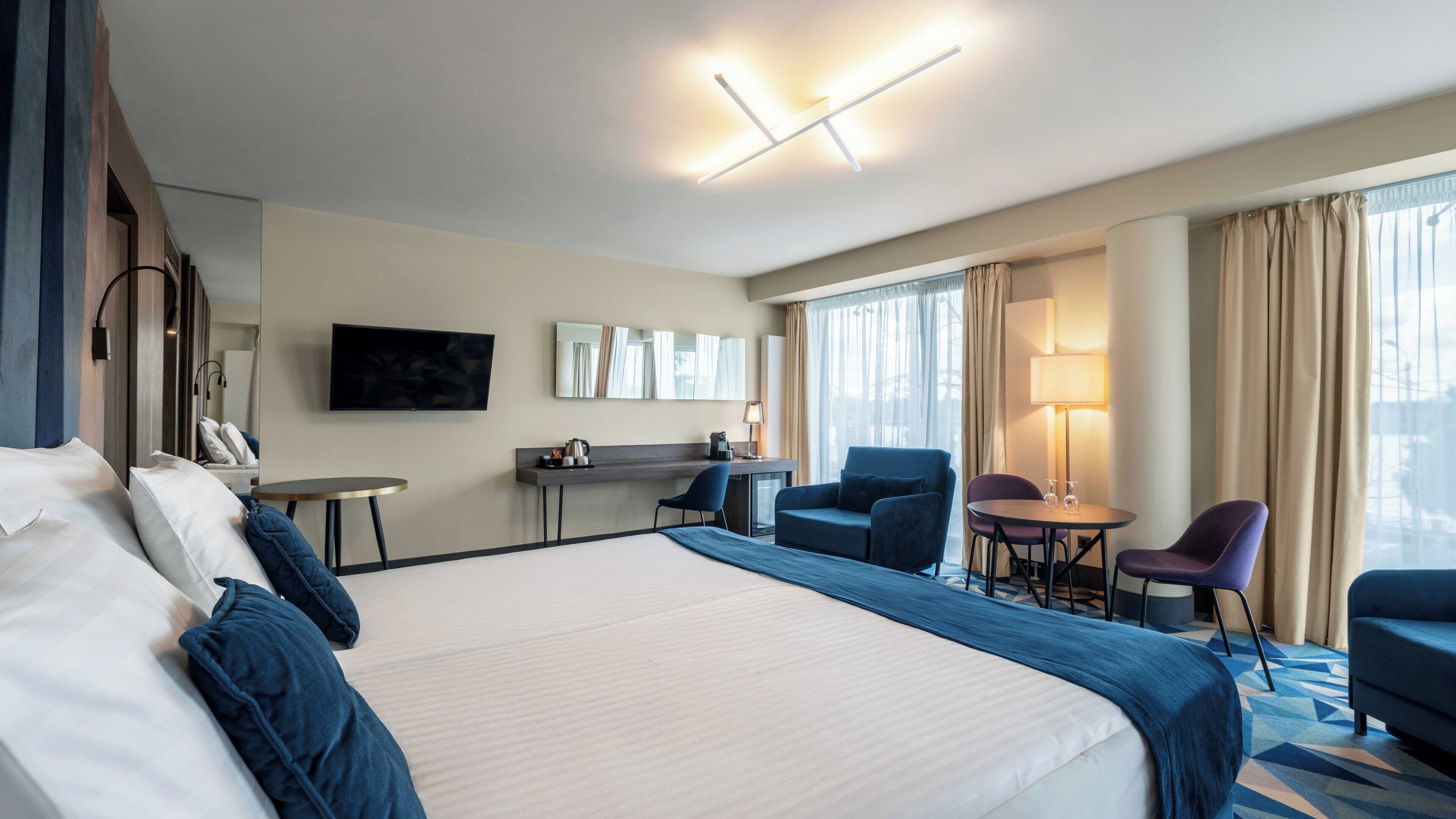 A hotel room with a large bed, a desk, a chair, a TV, and a window with curtains.
