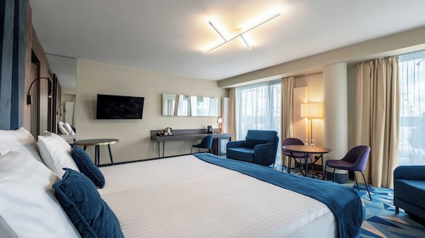 A hotel room with a large bed, a desk, a chair, a TV, and a window with curtains.